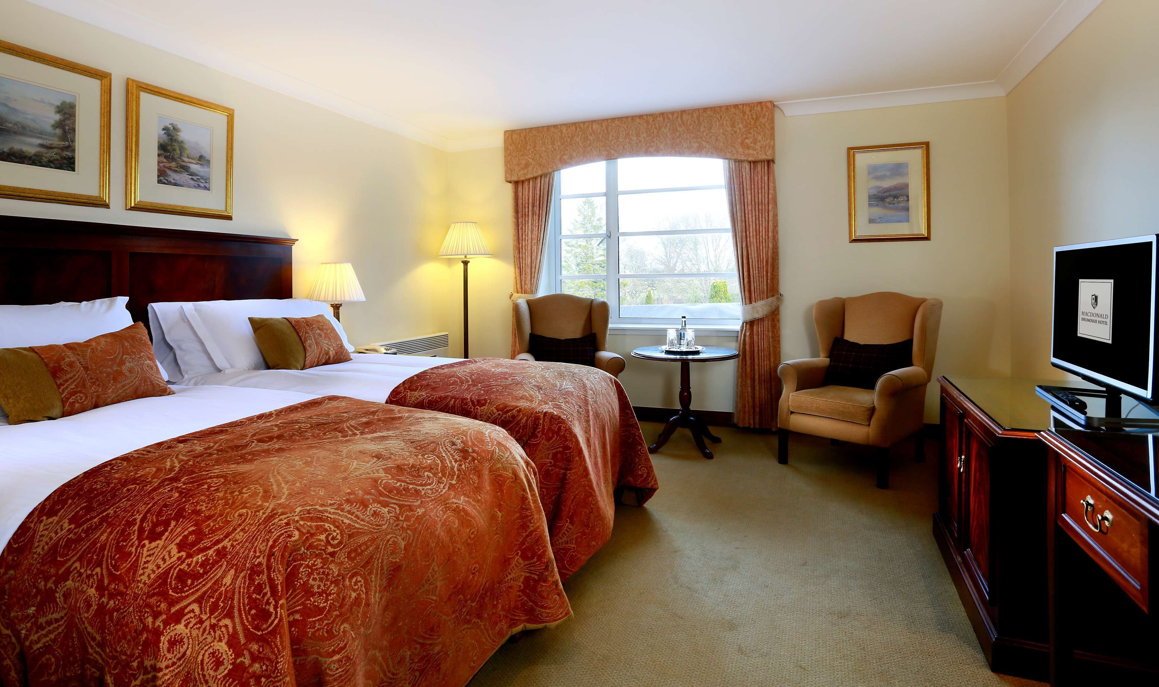 A hotel room with two beds, a desk, a chair, a TV, and a window with curtains.
