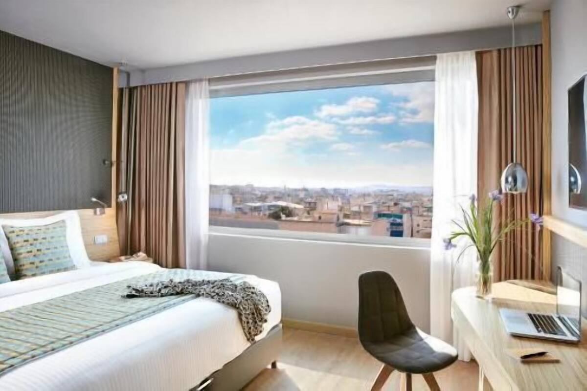 Una camera d'hotel con un letto, una scrivania con un portatile, una sedia e un'ampia finestra vista città.