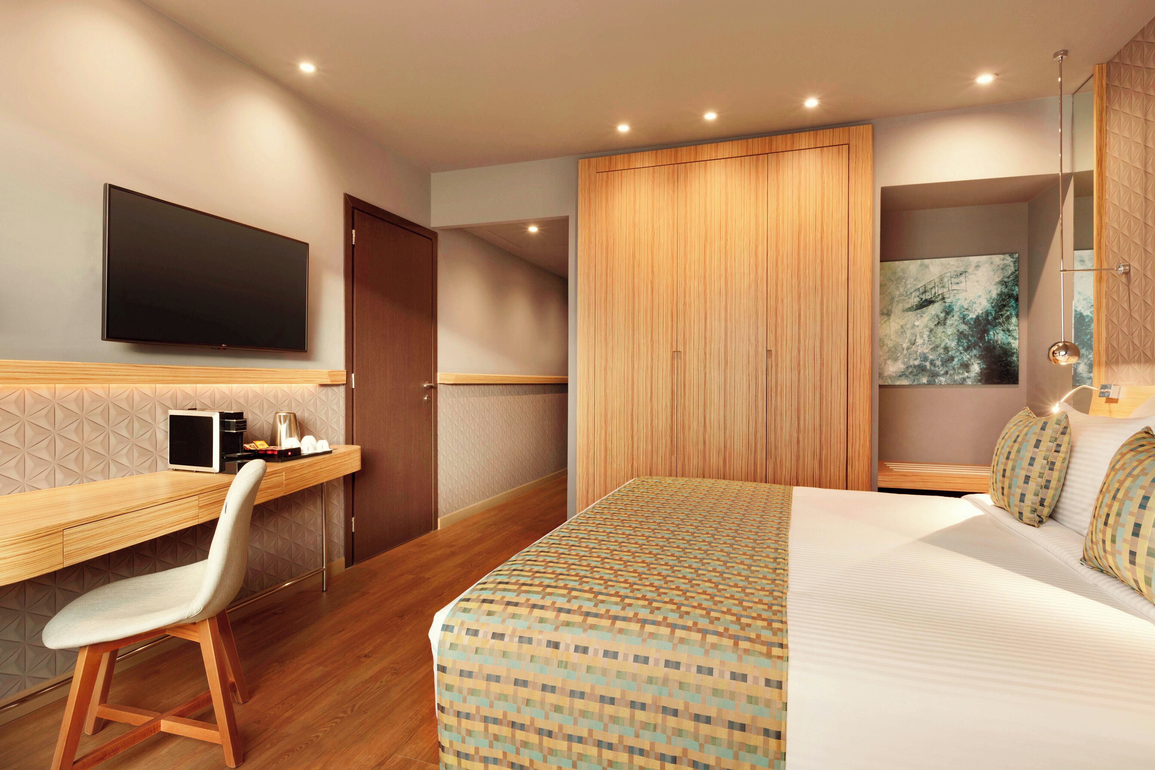 A modern hotel room with a large bed, a desk with a chair, a flat-screen TV, and a large window with a view of trees.