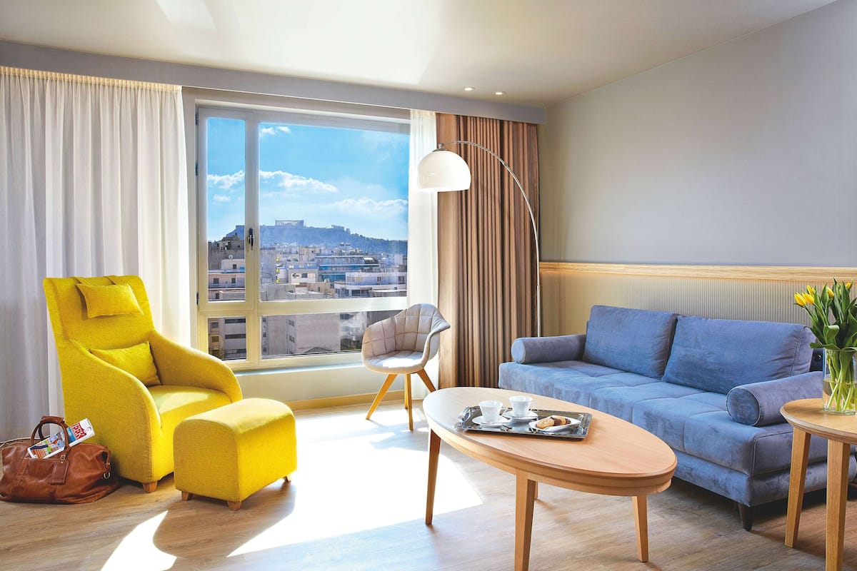 Camera d'albergo moderna con un'ampia finestra che offre vista sulla città, una poltrona gialla, un divano blu, un tavolino in legno e una lampada.