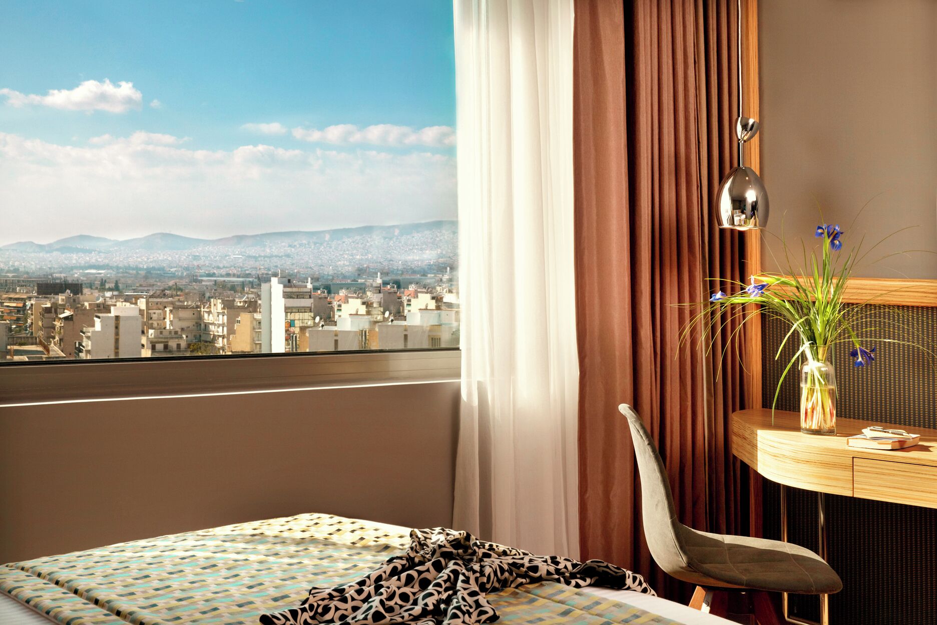 A hotel room with a bed, a chair, a desk with a vase of flowers, and a view of a cityscape.