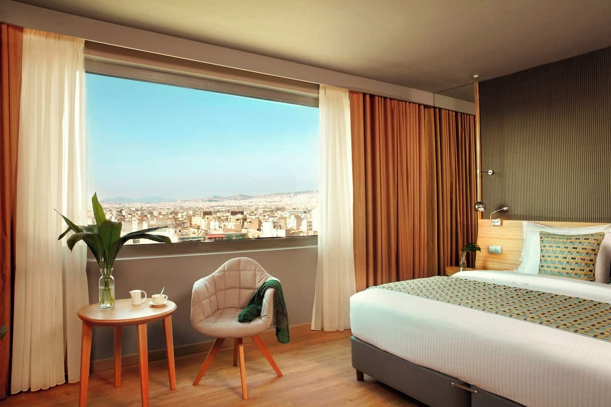 Una moderna camera d'albergo con un letto grande, un tavolino con una pianta e vista sulla città attraverso ampie finestre.