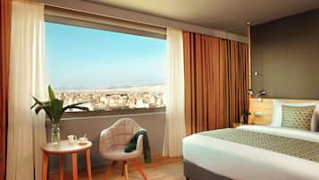 A modern hotel room with a large bed, a small table with a plant, and a view of a cityscape through large windows.