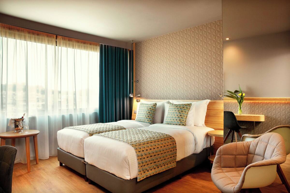 Camera d'albergo con un letto grande, un tavolino con una bottiglia e dei bicchieri, una sedia e uno scaffale a parete con un vaso.