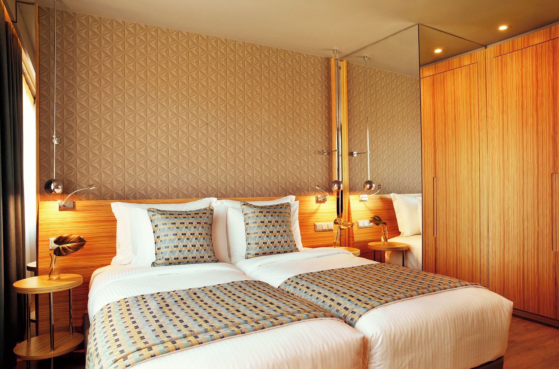 A double bed with a patterned bedspread, a wooden wardrobe, and a geometric patterned wall.
