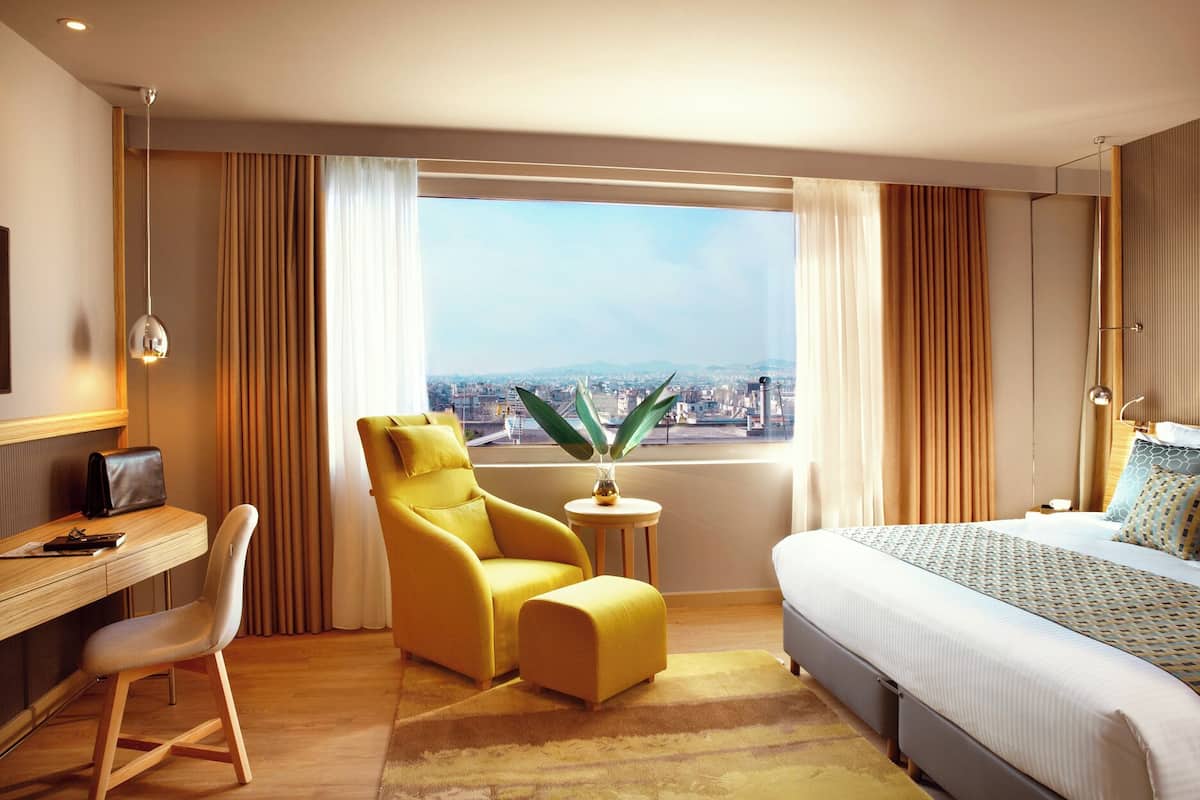 Una camera d'albergo con un letto, una scrivania, una sedia, un tavolino con una pianta e vista sulla città.