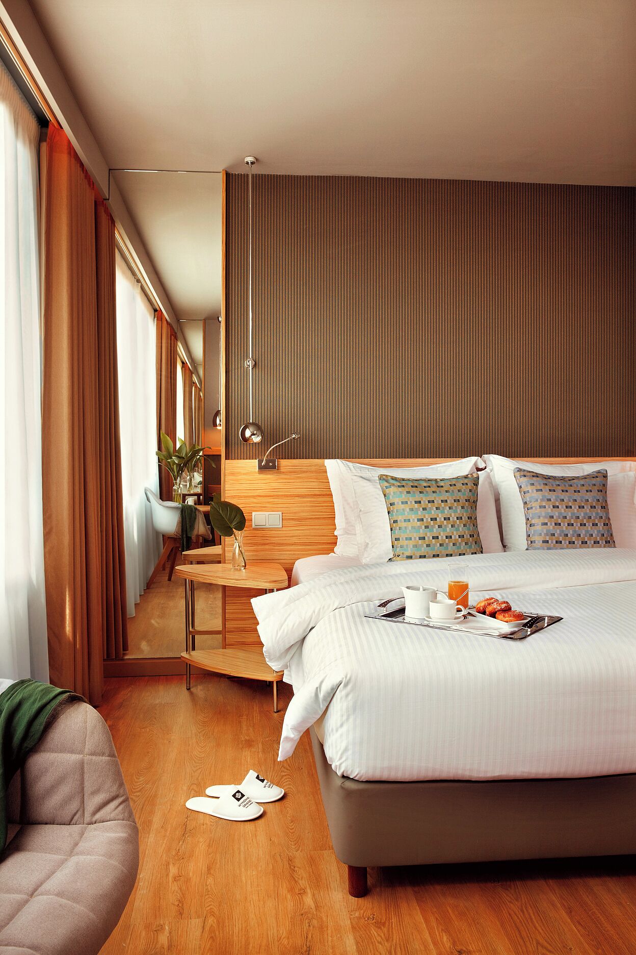 A modern hotel room with a large bed, a bedside table with a lamp, and a view of the city through large windows.