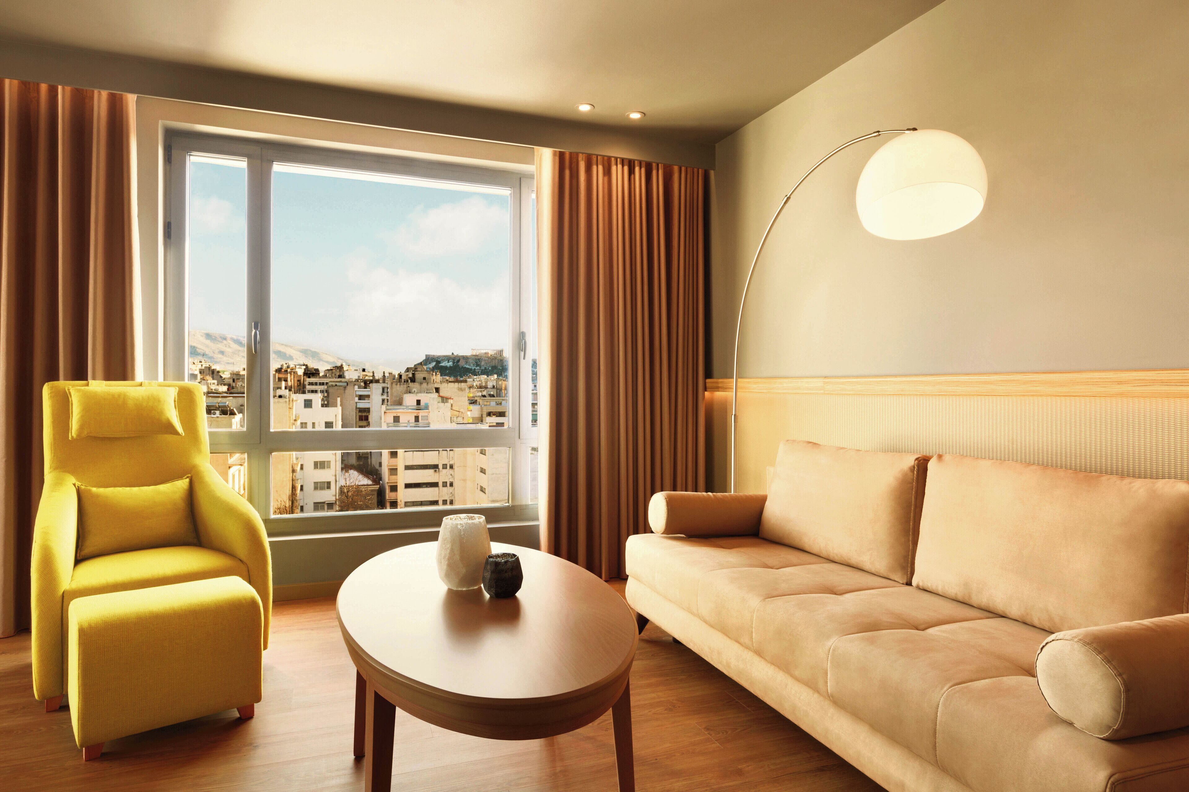 A modern hotel room with a yellow armchair, a beige sofa, a wooden coffee table, and a large window offering a city view.