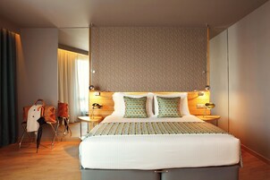 Quarto de hotel moderno com uma cama grande, cabeceira de madeira, duas mesas de cabeceira com abajures e vista para a cidade.
