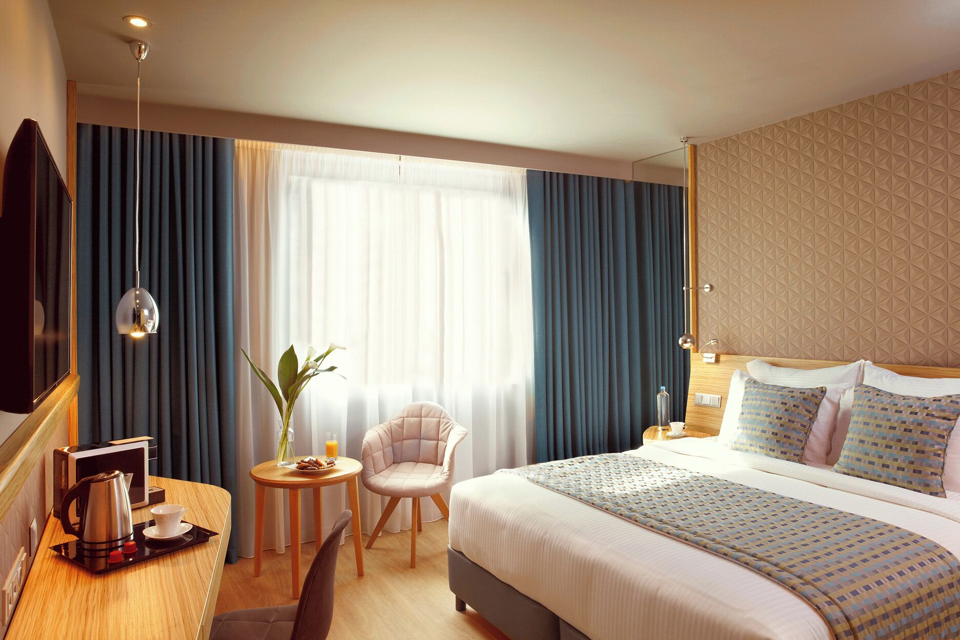A hotel room with a large bed, a small table with a kettle and cups, a chair, and a window with curtains.