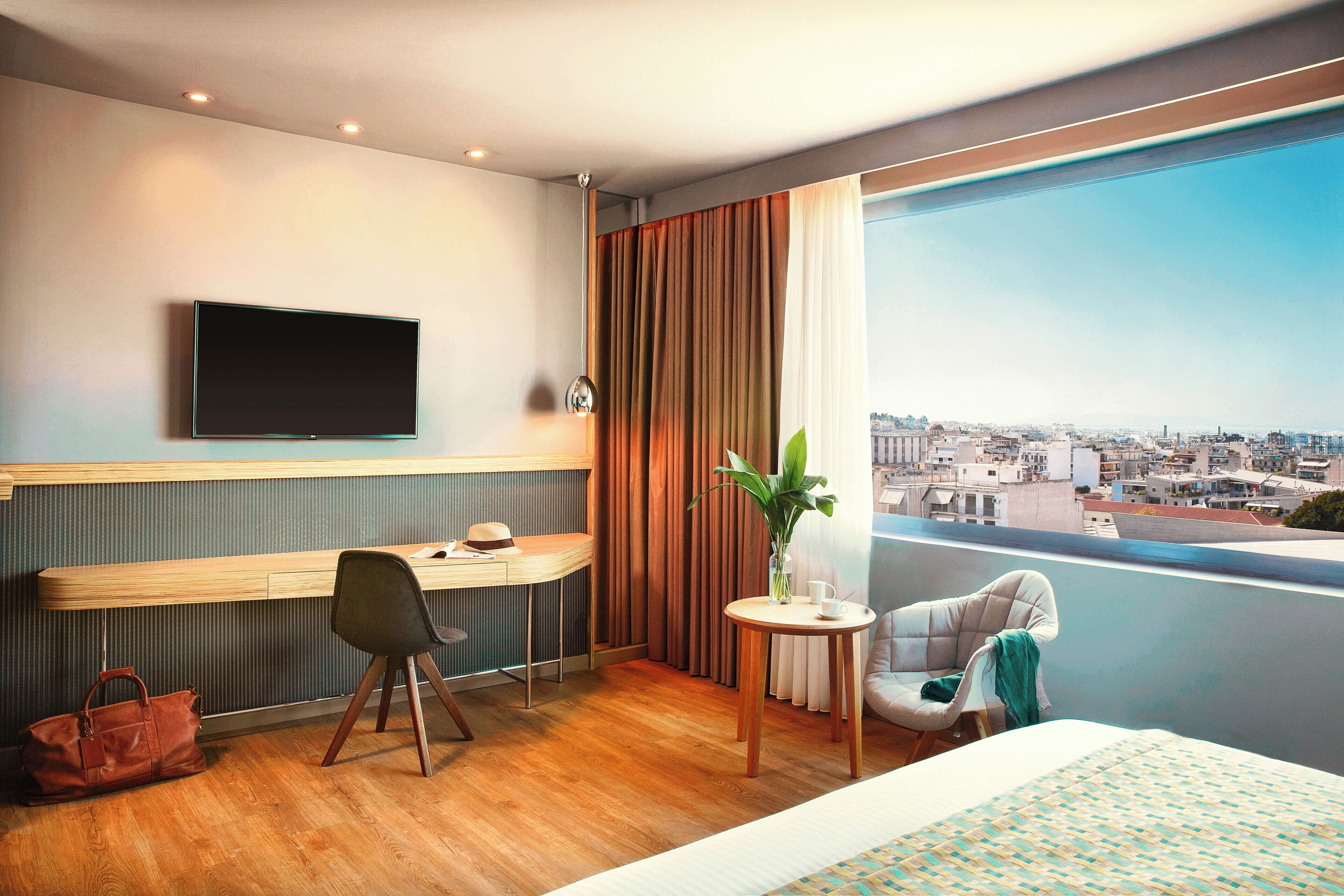 A hotel room with a desk, chair, TV, and a balcony with a city view.