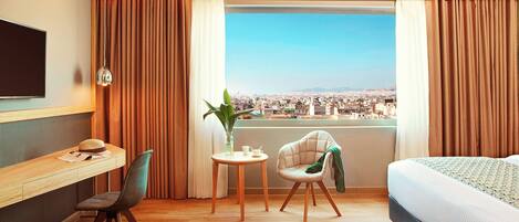 A hotel room with a bed, a desk with a chair, a small table with a plant, and a view of a cityscape through a large window.