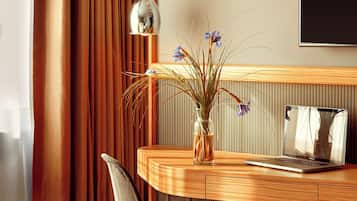 A hotel room with a bed, a desk with a laptop, a chair, a vase with flowers, and a TV on the wall.