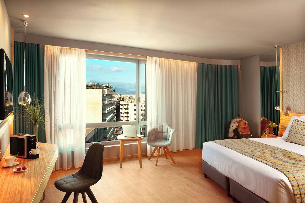 Camera d'albergo con un letto grande, una scrivania con un computer, due sedie, vista sulla città e un balcone.