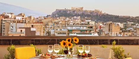 A rooftop dining area with a table set for four, a vase of sunflowers, and a view of a historic cityscape with a prominent hill and ancient ruins.