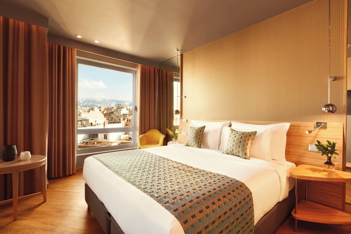 Una camera d'albergo con un letto grande, vista sulla città, un tavolino con un vaso e una testiera in legno.
