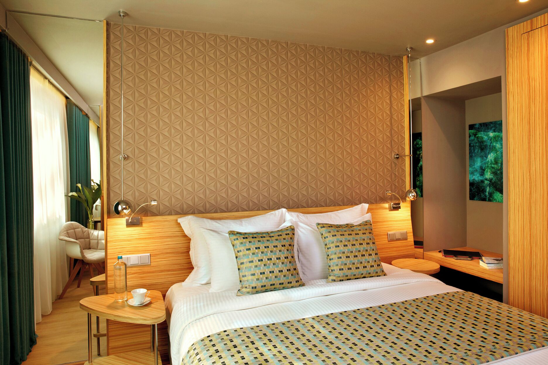 A modern hotel room with a geometric patterned headboard, a neatly made bed with patterned bedding, a small round table with a cup and a bottle, and a chair.