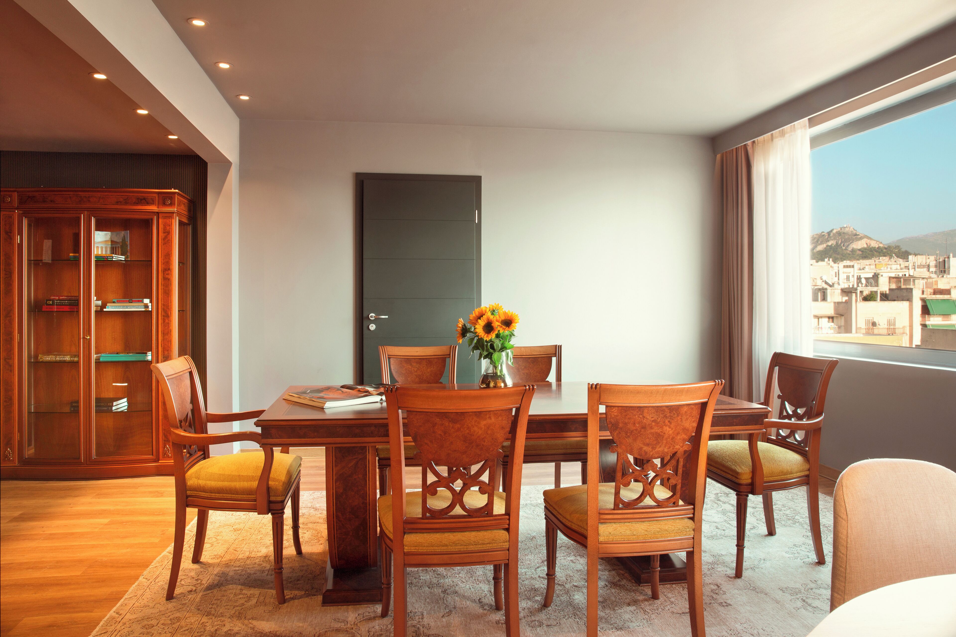 A dining area with a wooden table and chairs, a vase with flowers, and a view of a cityscape through a window.