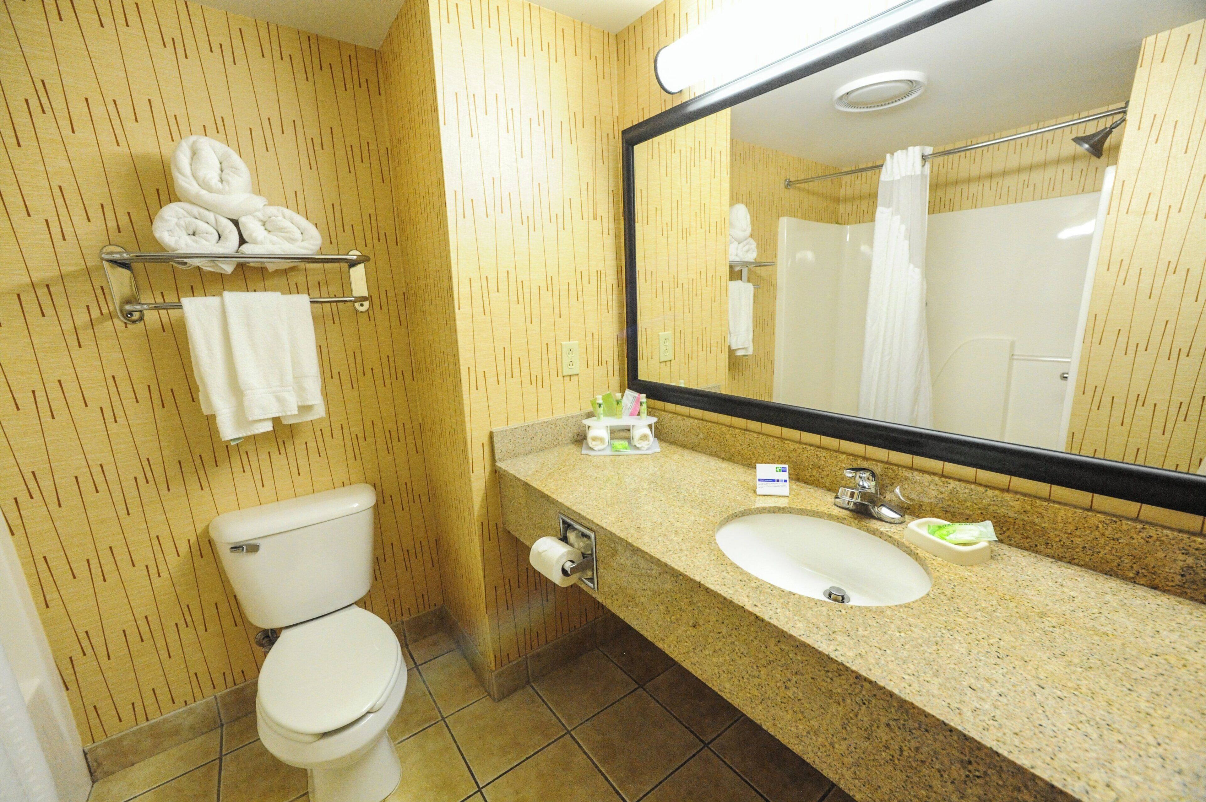Un baño con inodoro, lavamanos y espejo. Hay toallas en un perchero y un estante.