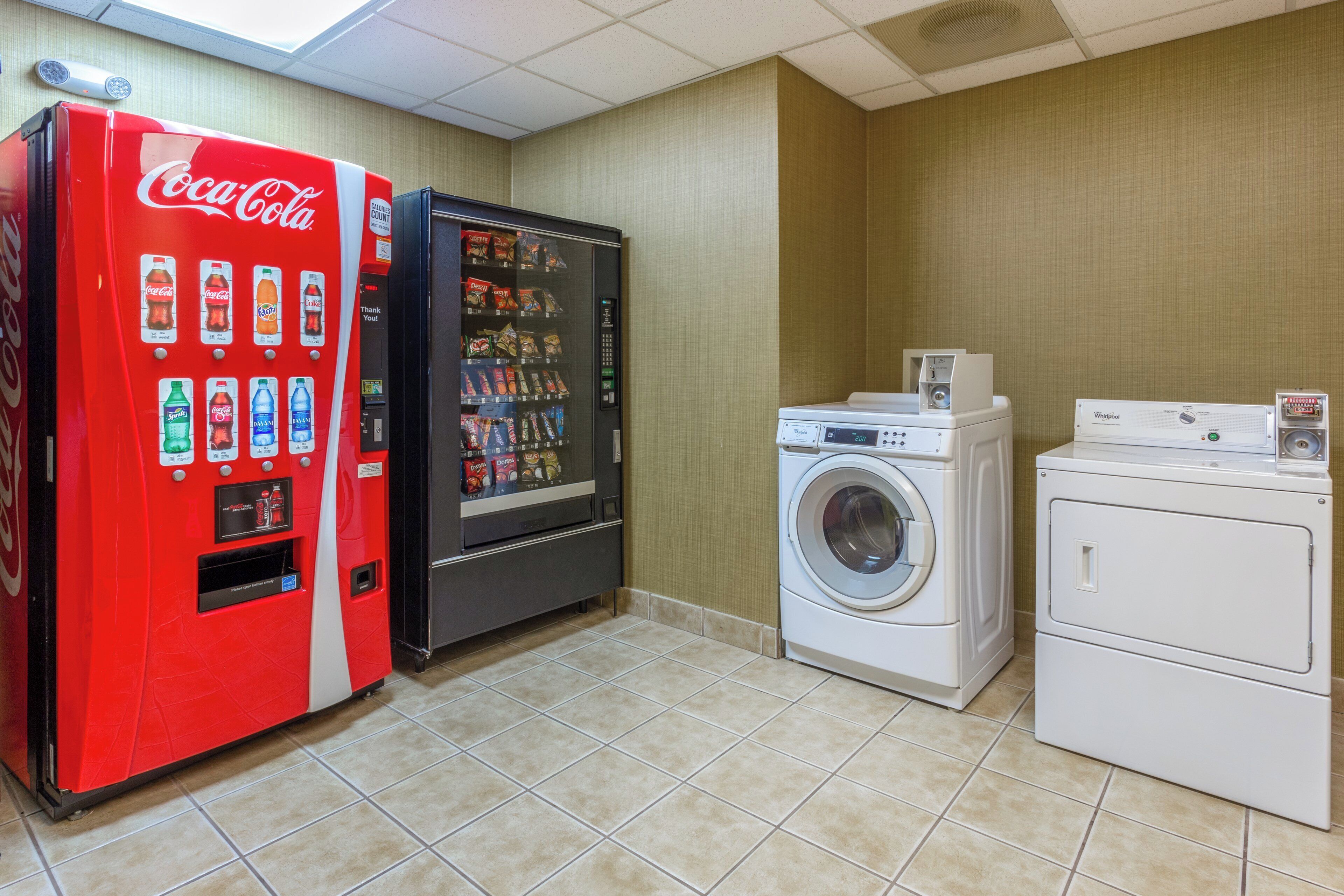 A room with a red Coca-Cola vending machine, a black refrigerator, a white washing machine, and a dryer.
