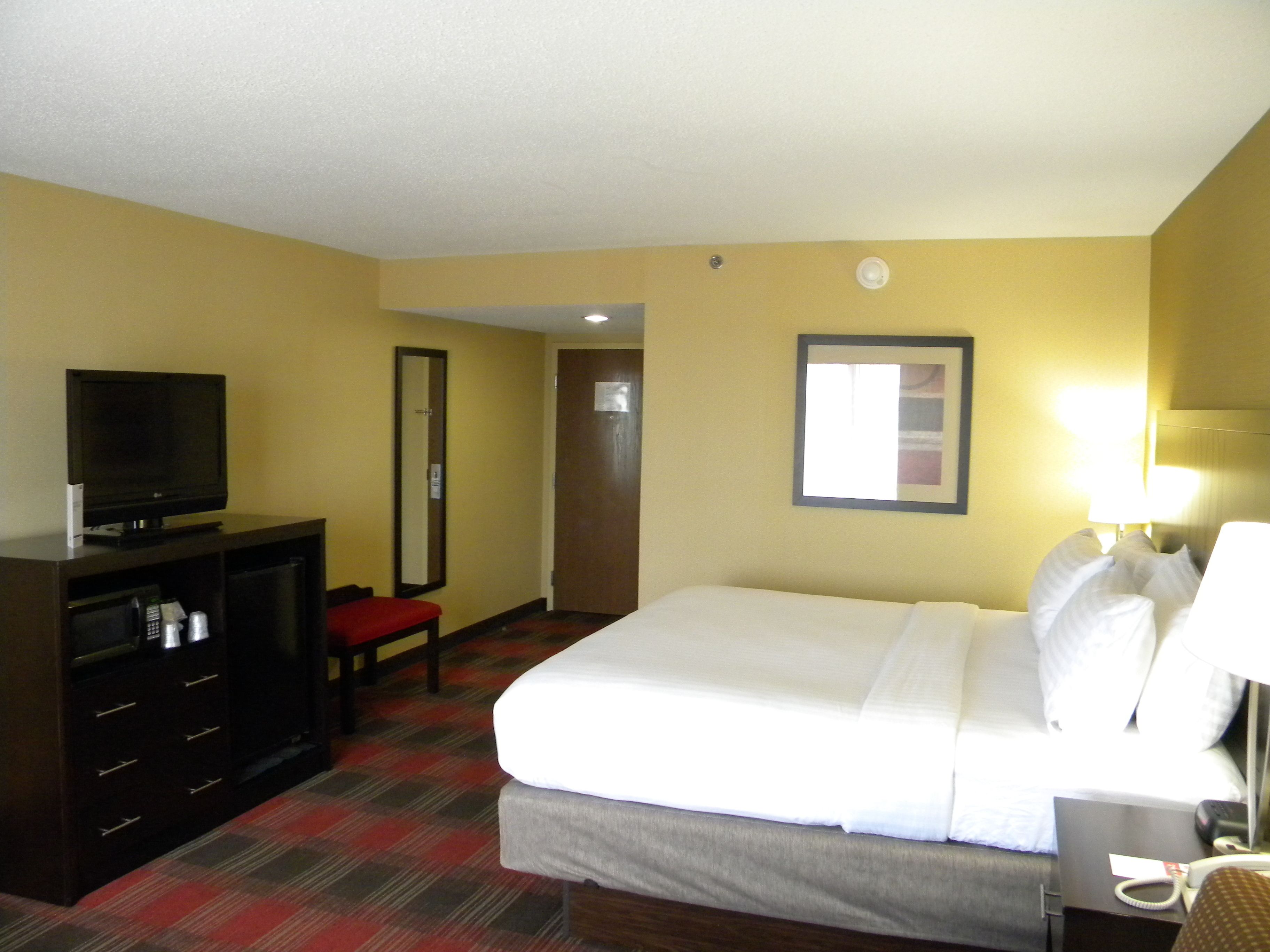 A hotel room with a bed, a television on a dresser, a desk with a lamp, and a mirror on the wall.