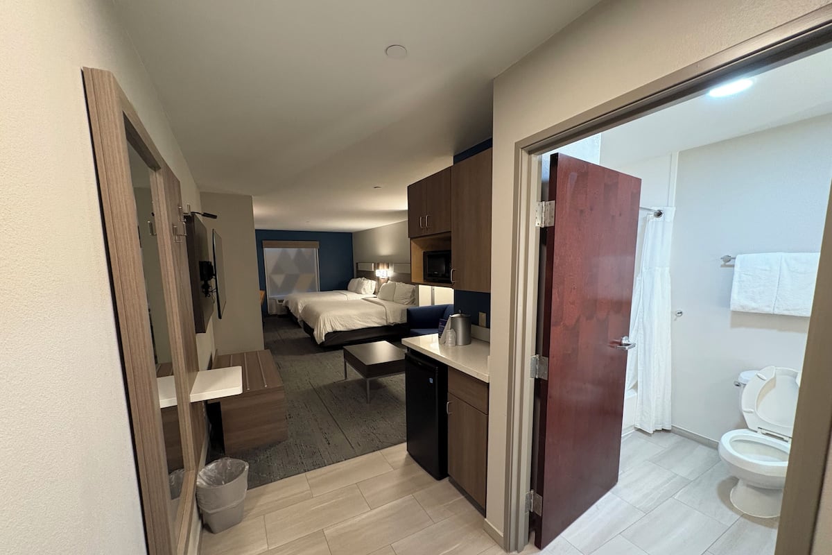 A hotel room with a bed, a desk, a chair, a small kitchenette, and a bathroom with a toilet and sink.