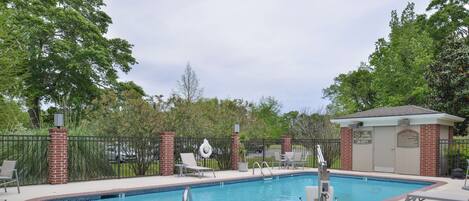 Una piscina rectangular con borde de azulejos, rodeada por un muro de ladrillo y una cerca de metal. Hay sillas de descanso y un pequeño edificio con un letrero en la cerca.