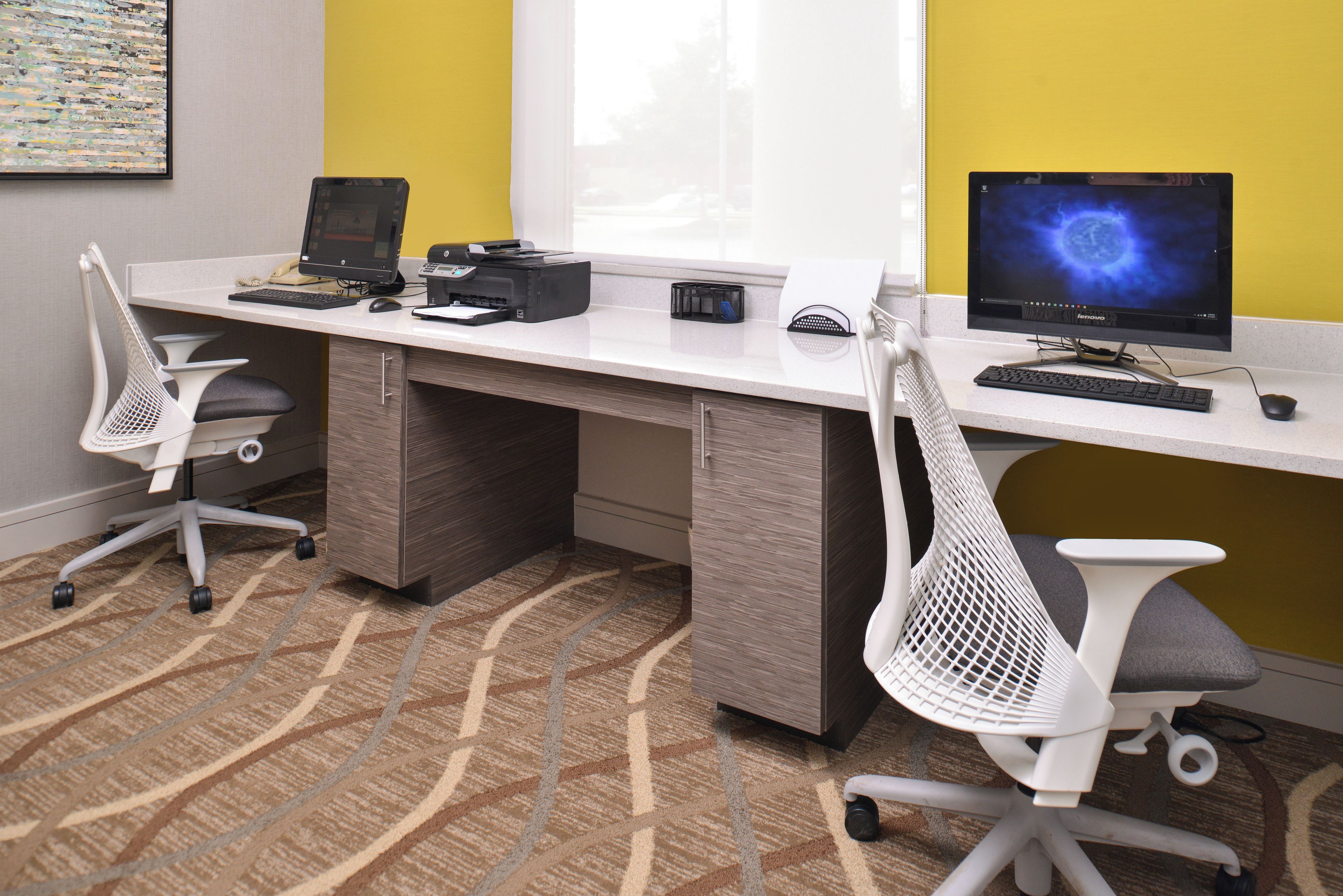 A modern office space with two desks, each equipped with a computer, keyboard, and mouse. There are two office chairs, one with a mesh backrest and the other with a solid backrest. The room has a patterned carpet and a window with blinds.