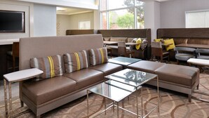 Um hall de hotel moderno com sofá de canto, mesas de vidro e uma área de estar com mesa alta.
