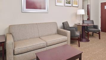 A hotel room with a beige sofa, two armchairs, a small wooden table, and a lamp.