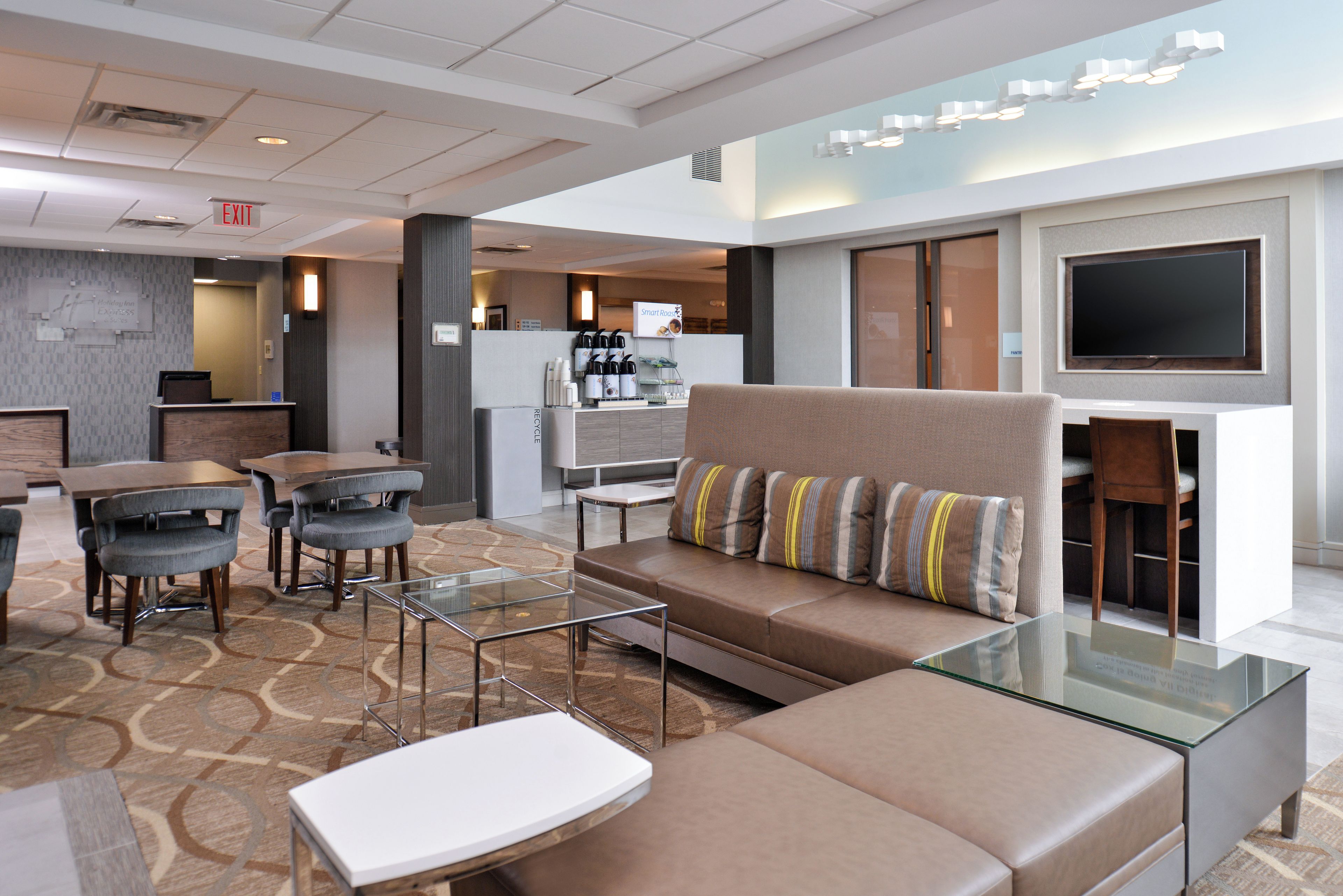 A modern hotel lobby with a seating area, a coffee station, and a flat-screen TV mounted on the wall.