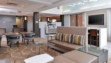 A modern hotel lobby with a seating area, a coffee station, and a flat-screen TV mounted on the wall.