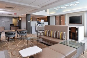 A modern hotel lobby with a seating area, a coffee station, and a flat-screen TV mounted on the wall.
