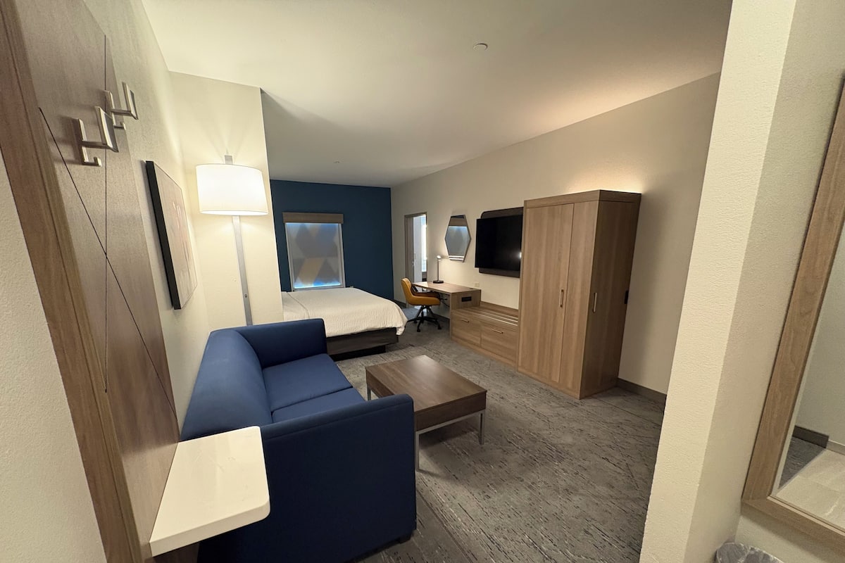 A compact hotel room with a bed, a blue sofa, a desk with a chair, a TV, and a wardrobe.