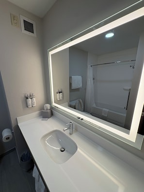 A modern bathroom with a large mirror, white sink, and a shower visible in the reflection. - Holiday Inn Express Hotel & Suites Lafayette by IHG (Lafayette)