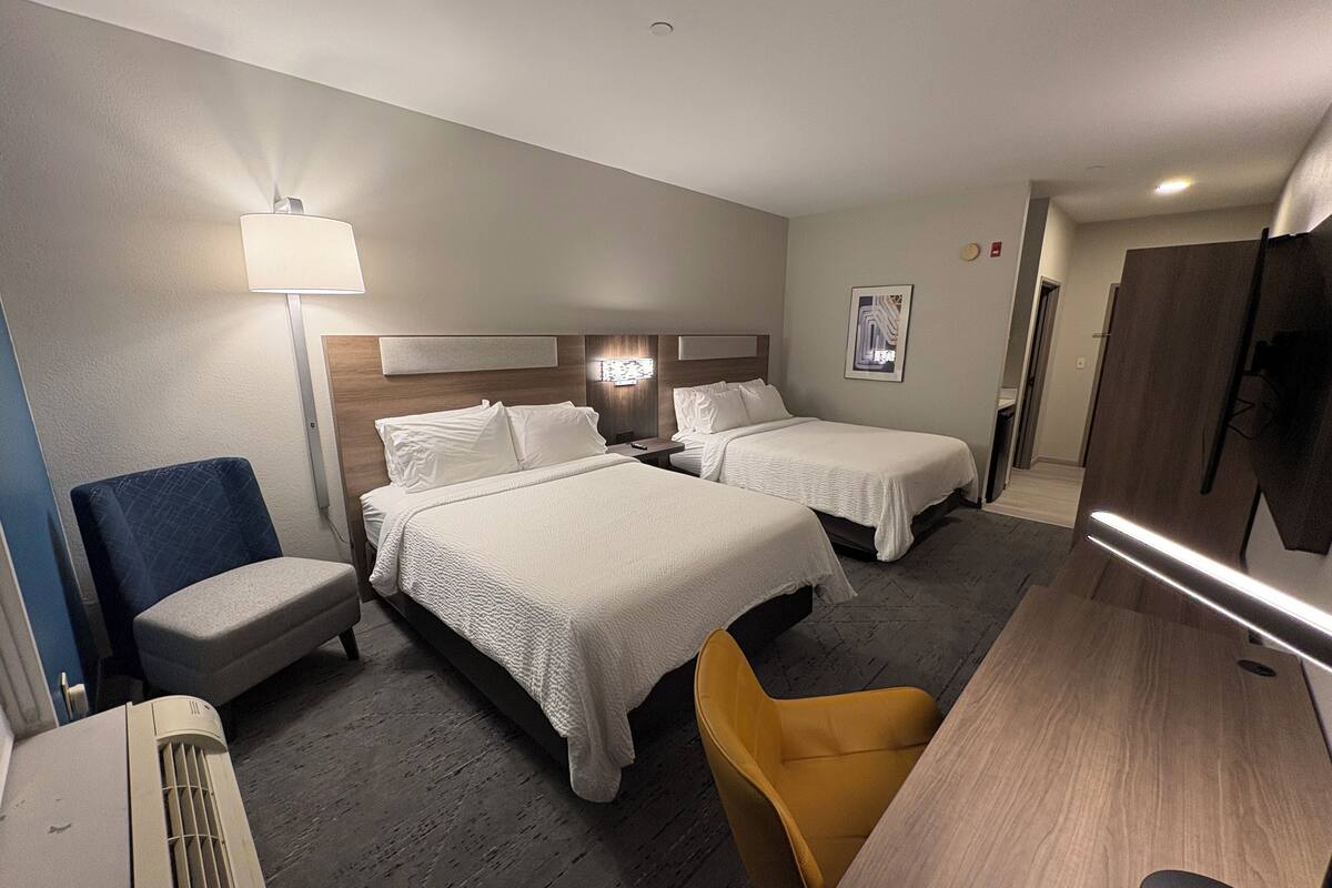 A hotel room with two beds, a desk, a chair, a lamp, and a TV.