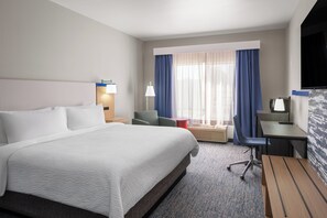 Et hotelværelse med en stor seng, et skrivebord, en stol, et fjernsyn og et vindue med gardiner.