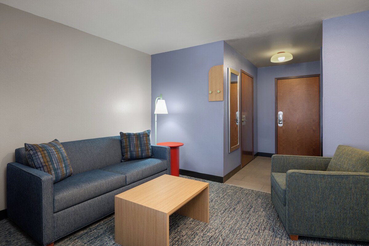 A modern hotel room with a grey sofa, a wooden coffee table, a red side table, and a green armchair.