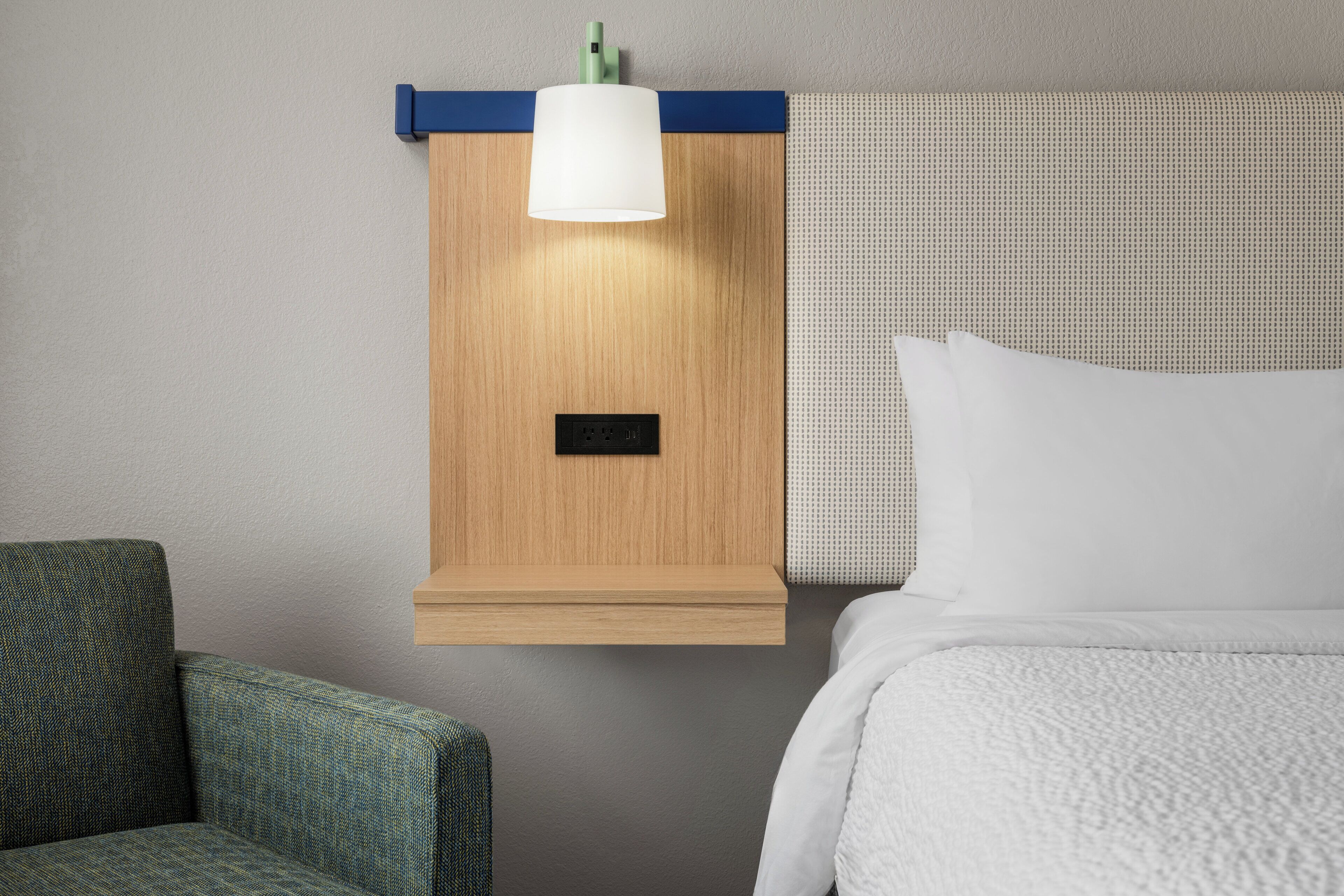 A hotel room with a wooden nightstand, a lamp, a bed with white bedding, and a green upholstered armchair.