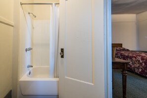 A bathroom with a bathtub and shower, a door leading to a bedroom with a bed and a wooden headboard.