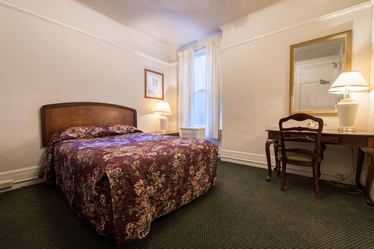 Une chambre d’hôtel avec un lit, un bureau, une chaise, une lampe, une fenêtre avec des rideaux et un miroir au mur.