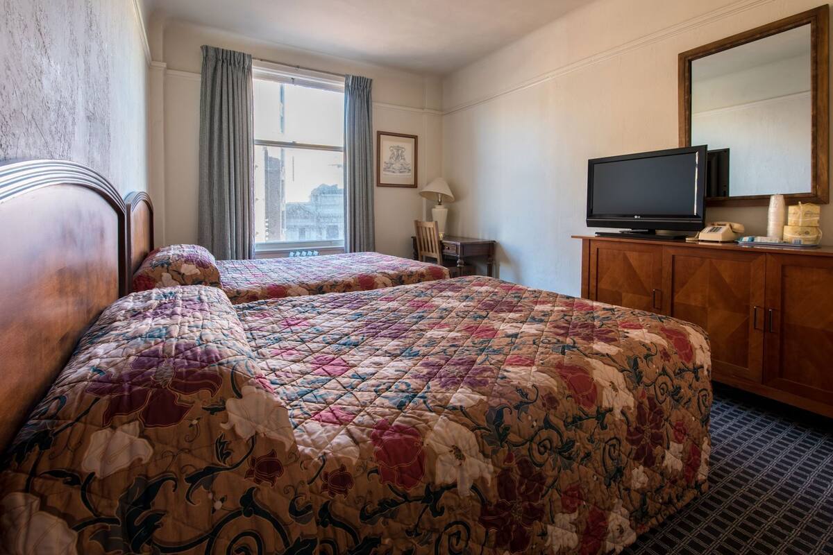 Une chambre d’hôtel avec deux lits, une télévision, un bureau et une fenêtre avec des rideaux.