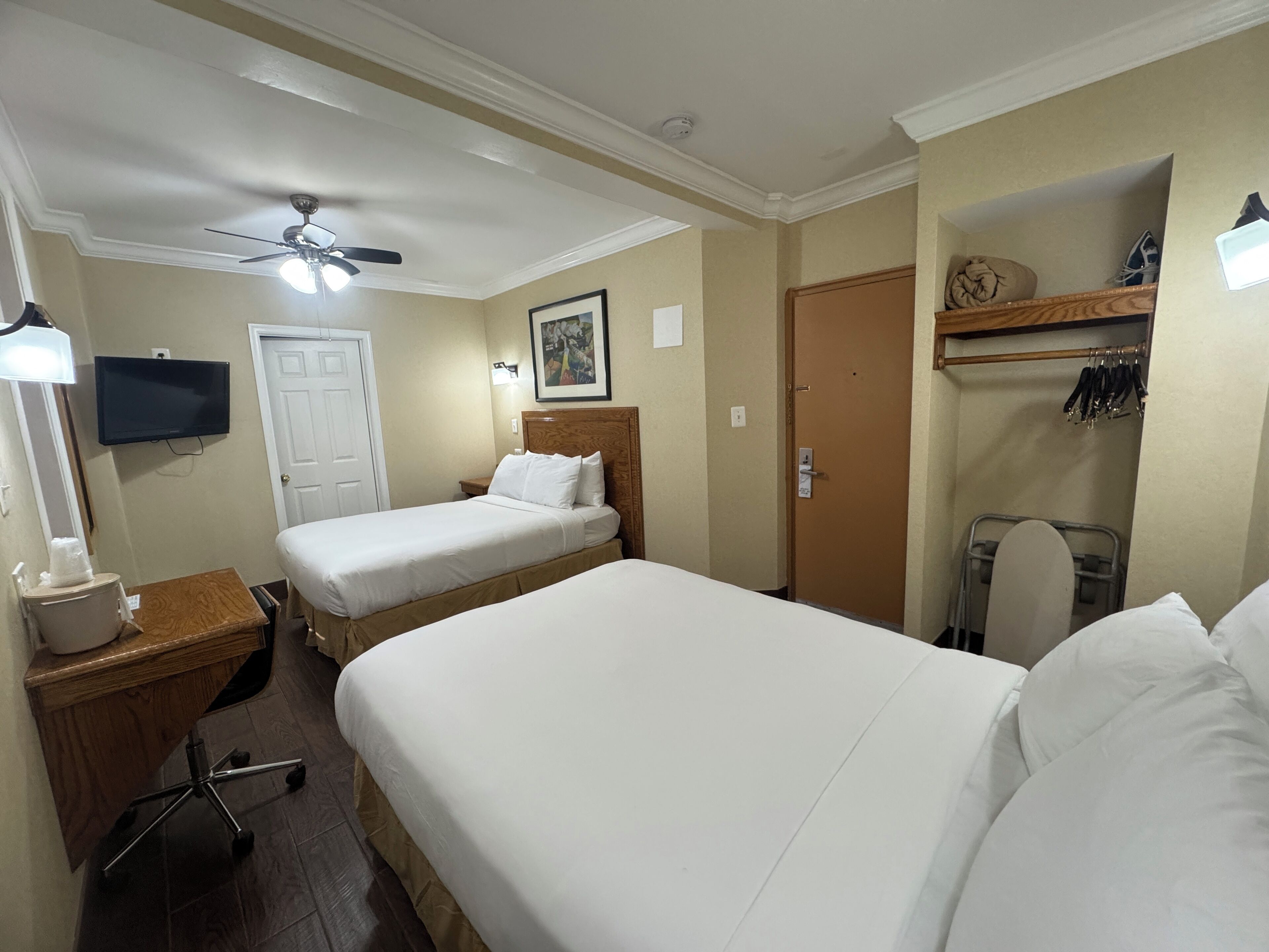 A hotel room with two beds, a ceiling fan, a TV, a wooden dresser, a desk with a chair, and a coat rack.
