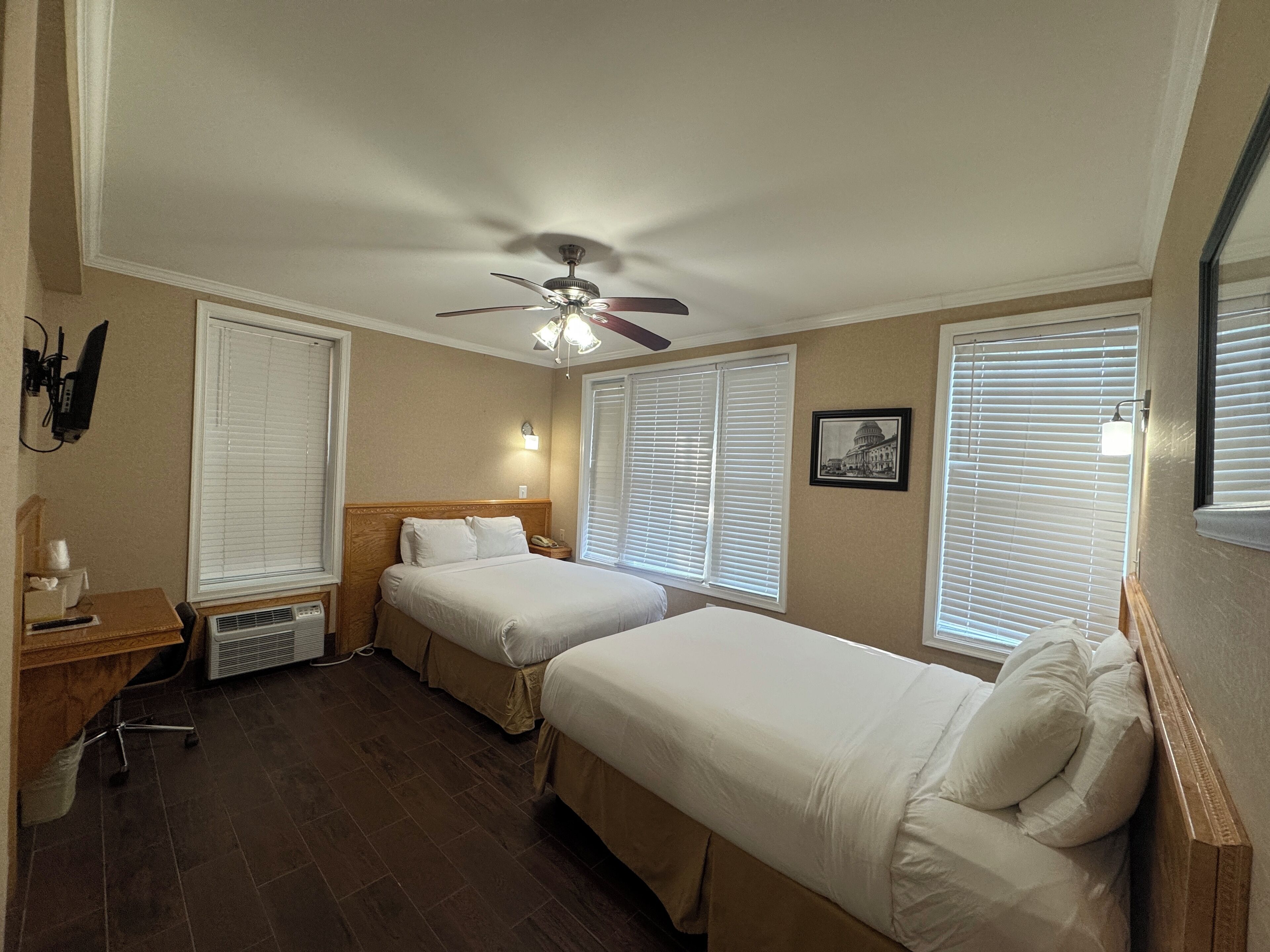 Habitación de hotel con dos camas individuales, ventilador de techo, escritorio con silla y ventana con persianas.