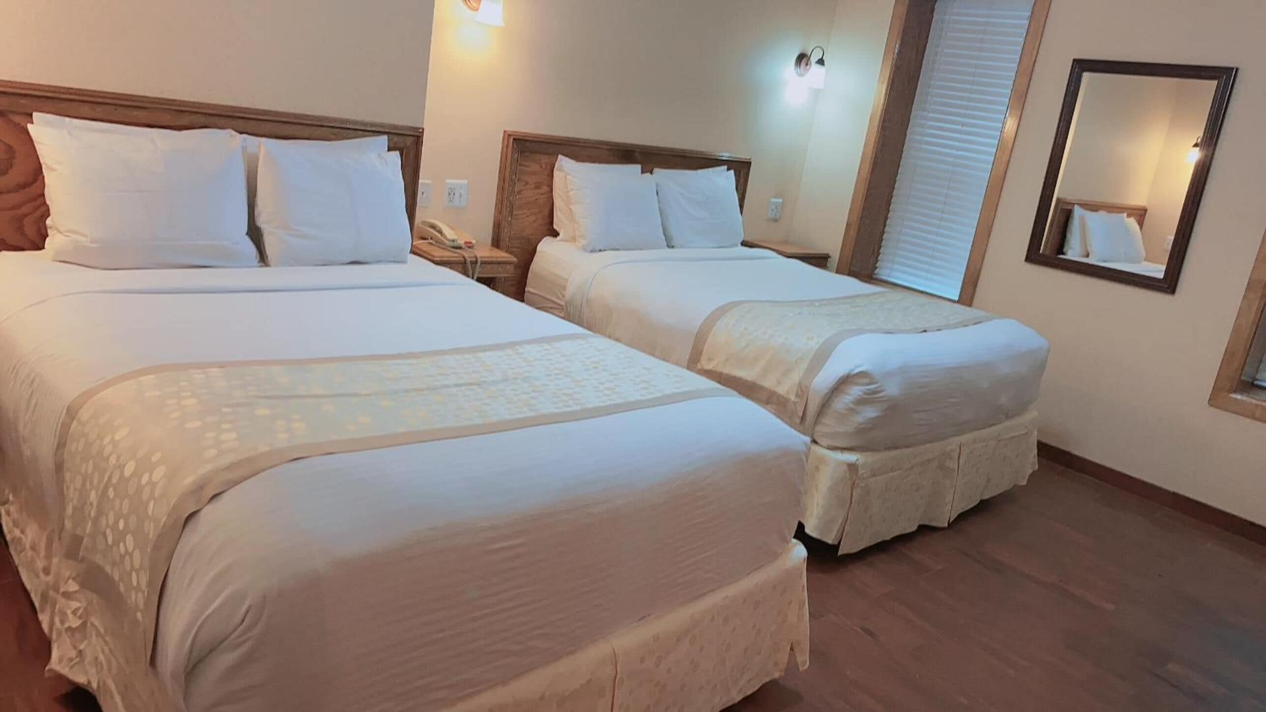 Habitación de hotel con dos camas, cada una con ropa blanca y cabeceros, una mesita de noche con lámpara y un espejo en la pared.