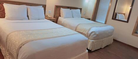 Ein Hotelzimmer mit zwei Betten, jeweils mit weißer Bettwäsche und Kopfteilen, einem Nachttisch mit Lampe sowie einem Spiegel an der Wand.