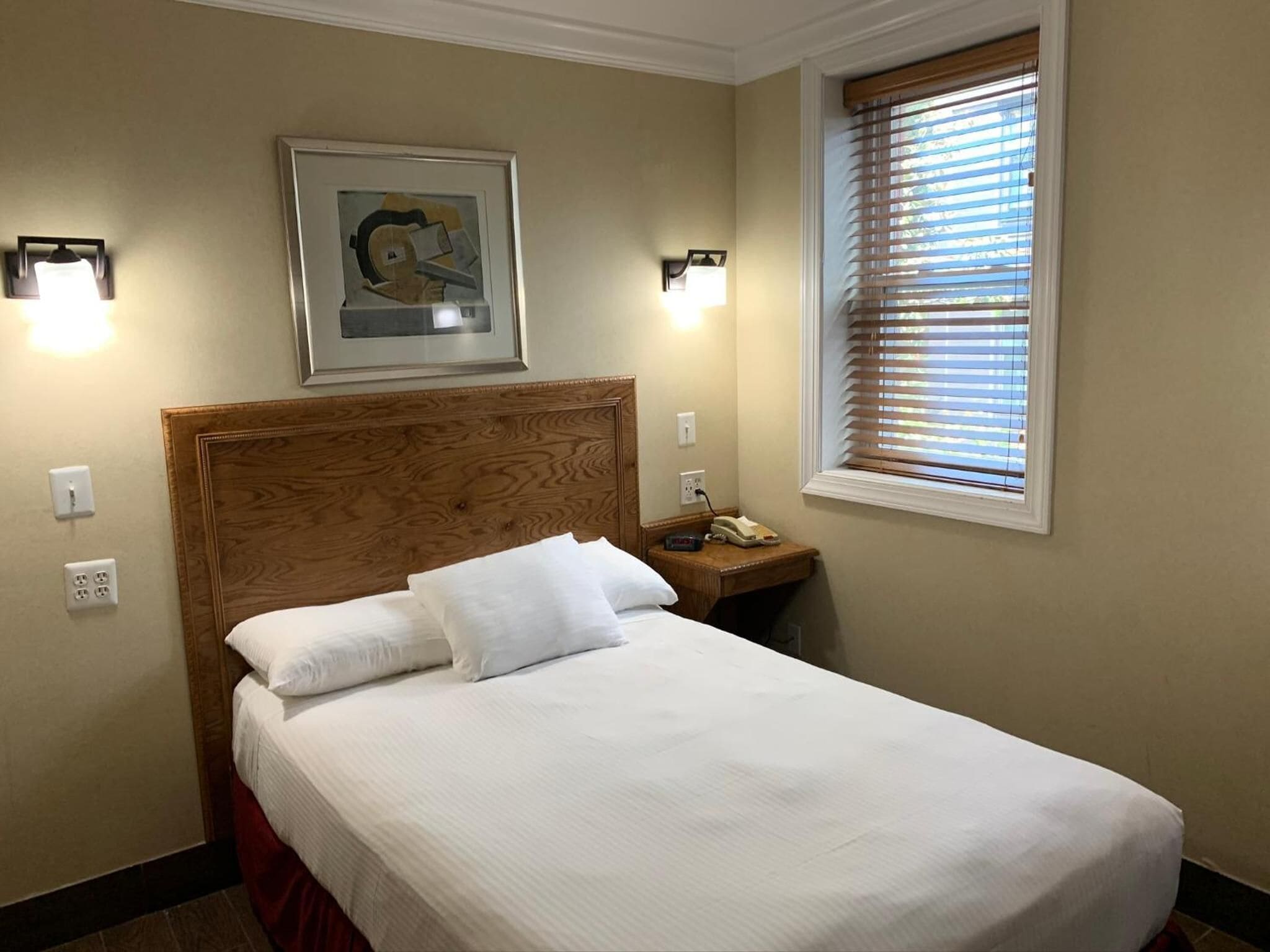 A hotel room with a bed, a nightstand, a framed picture, and a window with blinds.