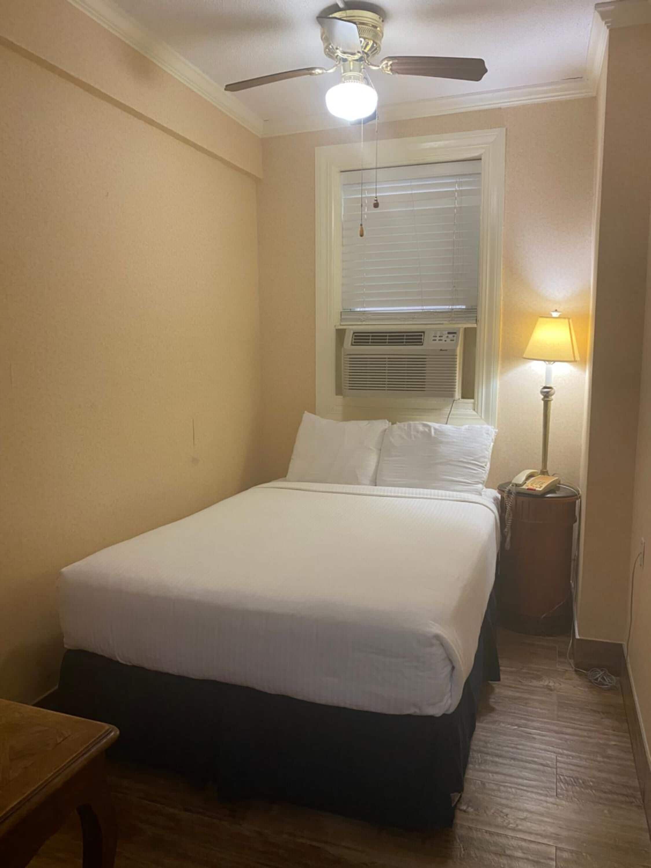 A single bed with a headboard, a bedside table, a lamp, and a ceiling fan in a room with beige walls and a wooden floor.
