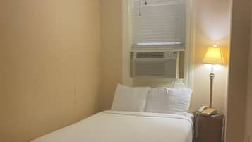 A single bed with a headboard, a bedside table, a lamp, and a ceiling fan in a room with beige walls and a wooden floor.