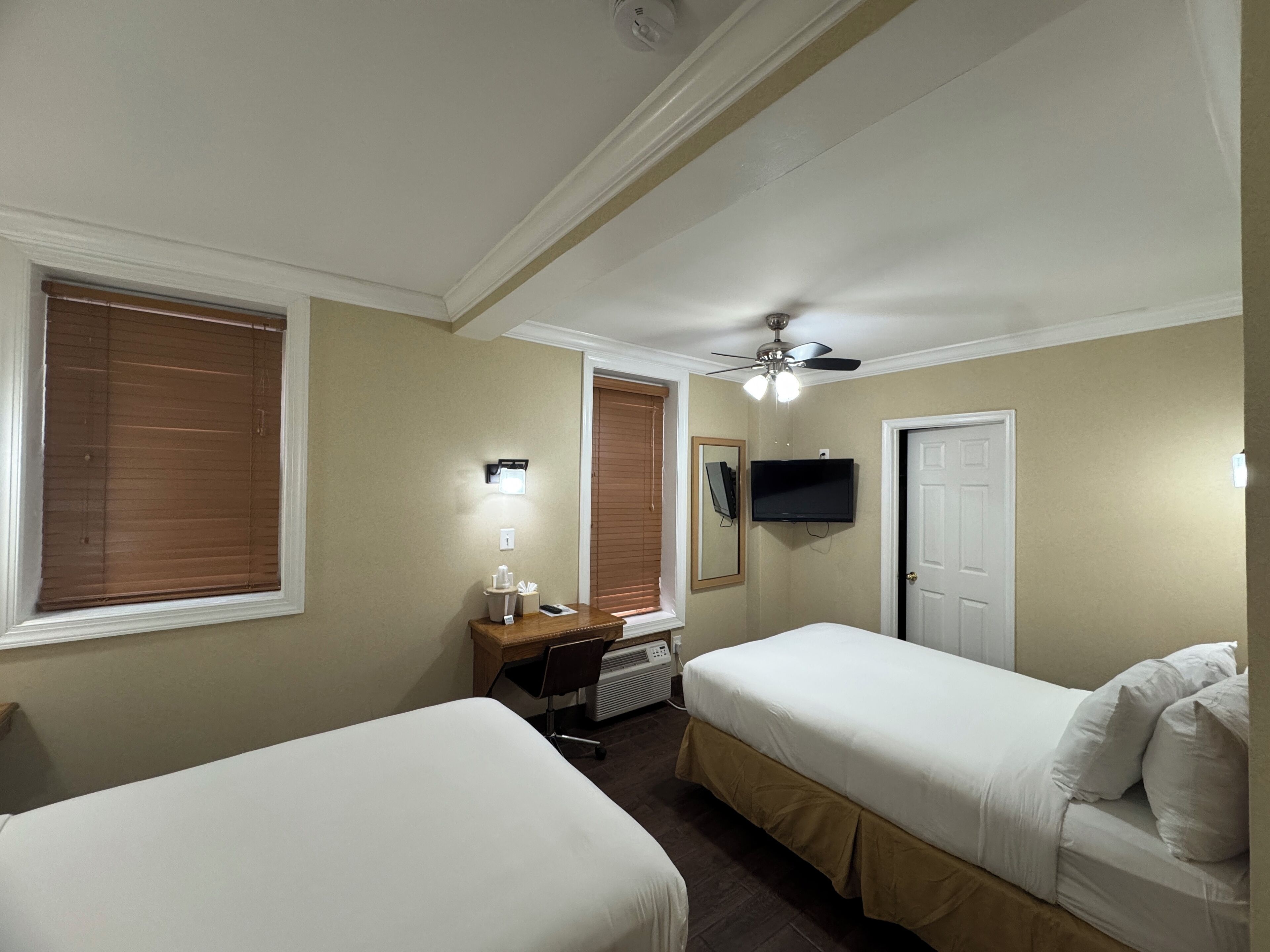 A hotel room with two beds, a desk with a chair, a ceiling fan, a TV mounted on the wall, and a door.